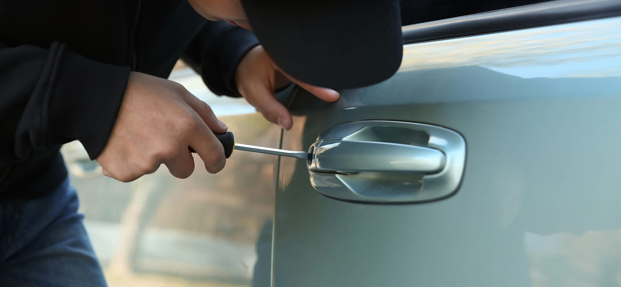 A person wearing a cap and jacket is attempting to break into a car using a screwdriver at the door handle. The car appears to be silver. The individuals face is not visible.