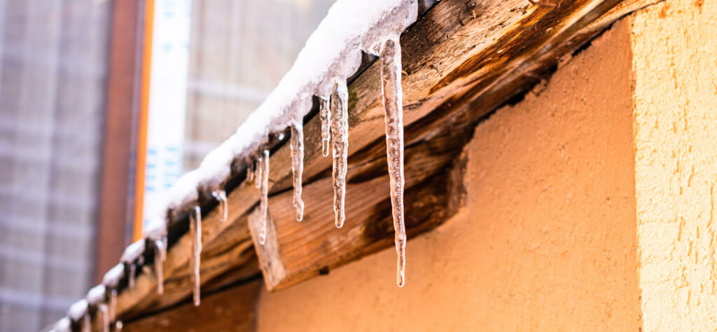 Icicles hang from the edge of a snow-covered wooden roof, glistening in the sunlight. The warm tones of the buildings wall contrast with the cold, wintry scene above.