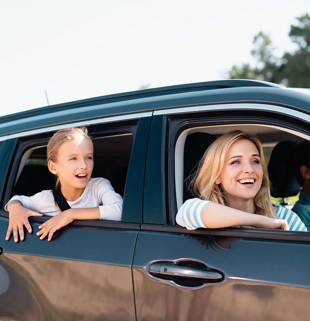 A woman and a child are smiling as they look out of the windows of their parked black car, secure in their travels. The woman is in the front seat, and the child sits joyfully in the back. With auto insurance peace of mind, they enjoy a day framed by a clear sky and lush greenery.