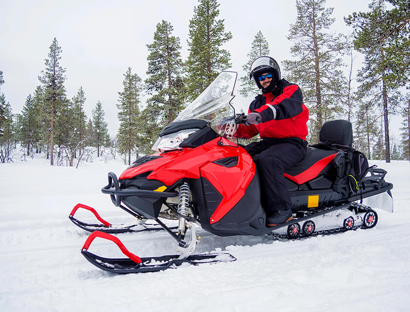 A person wearing a red and black jacket and helmet is riding a red snowmobile across snowy terrain, surrounded by snow-covered trees. With a smile, they enjoy the ride in the wintry landscape, knowing their auto insurance has them covered for any unexpected adventures.