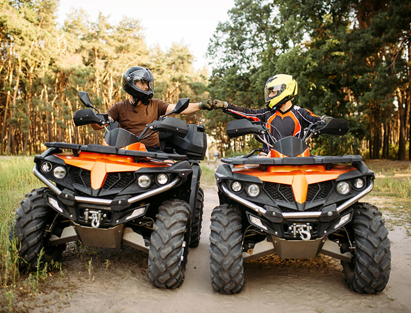 Two people wearing helmets fist-bump while seated on orange and black all-terrain vehicles (ATVs), their adventurous spirits roaming free on a dirt path. Surrounded by a forest of tall trees under a clear sky, theyre glad they remembered to check their auto insurance before setting off.