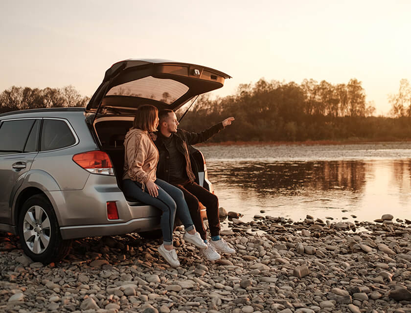 A couple relaxes on the open tailgate of a silver SUV parked by a pebble-covered riverbank, enjoying the serenity. As the sun sets behind them, they point toward the water, assured in their adventure and their choice of reliable auto insurance for peace of mind.