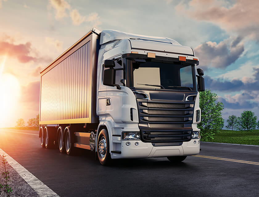 A large white semi-truck with a trailer drives on a highway at sunset, showcasing the importance of reliable auto insurance. The sky is partly cloudy, casting an orange glow on the road flanked by green trees and grass.