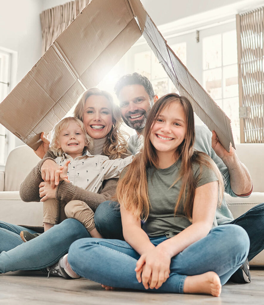 A smiling family of four sits on the floor in a bright room, embodying strong roots. The parents hold a cardboard piece shaped like a roof over their heads, while a young girl sits cross-legged in front and a small child is cradled by the mother. Light streams through a window behind them.