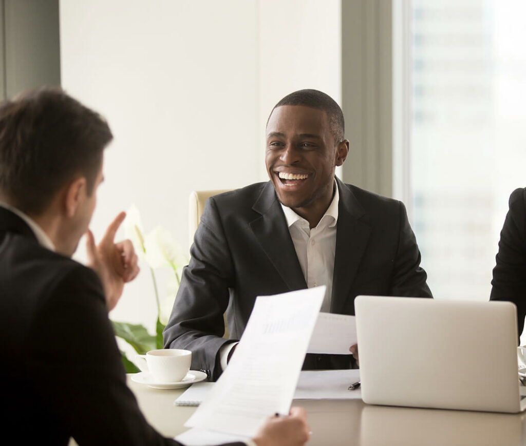 A person in a suit smiles across the table during a meeting on commercial insurance. Another participant, partially visible, gestures animatedly while discussing details. A laptop and coffee cup rest on the table, with bright windows providing a vibrant backdrop.