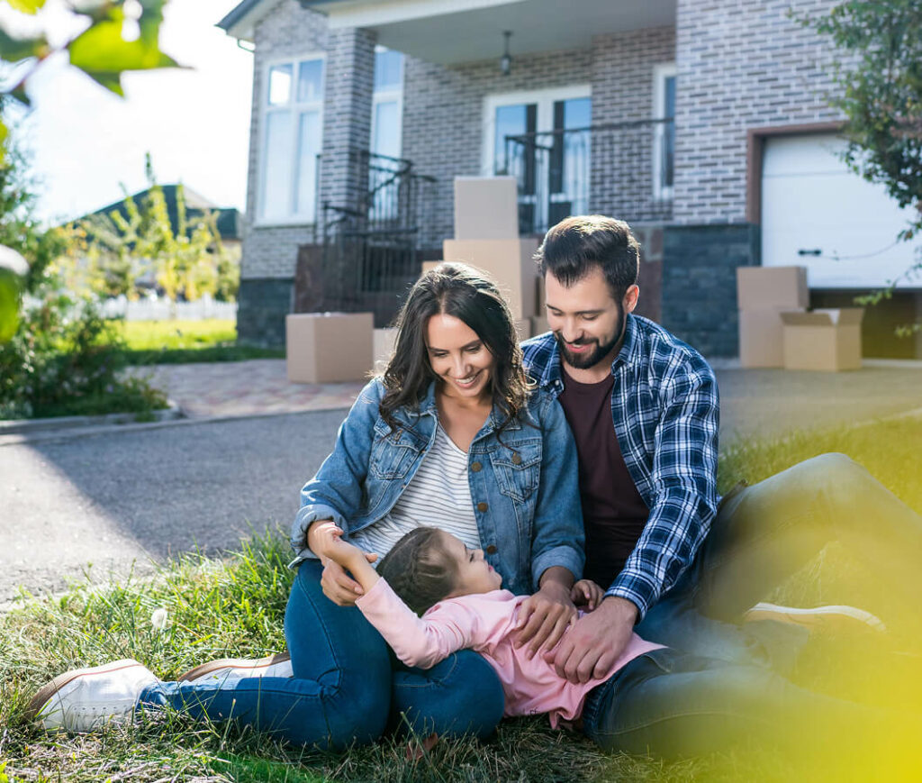 A family of three sits on the grass in front of a house with moving boxes nearby, their new haven secured by residential insurance. The mother and father smile warmly at their child, who is lying across their laps, enjoying a sunny day.