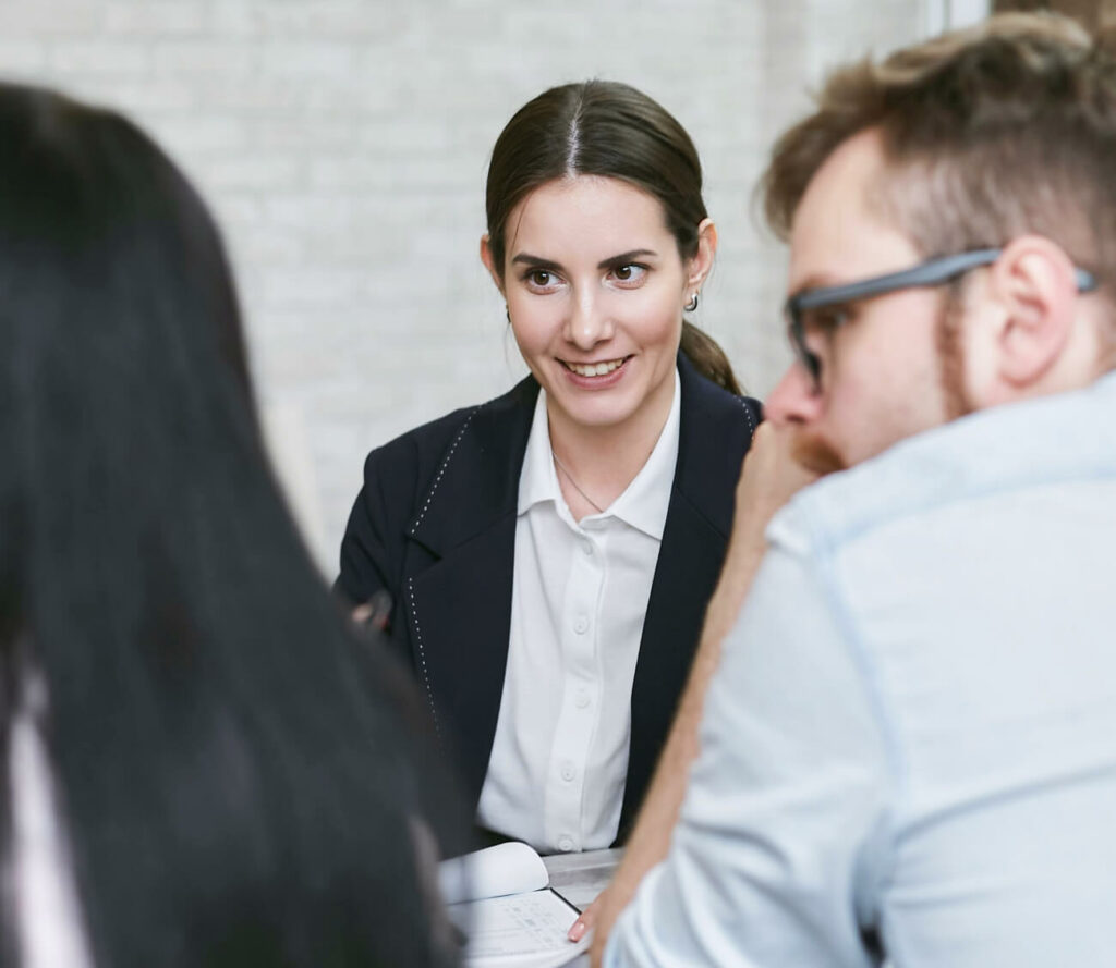 A woman in a white shirt and dark blazer is smiling while sitting at a table with two other people in an office setting. She appears to be engaged in conversation. The background features a light-colored brick wall.