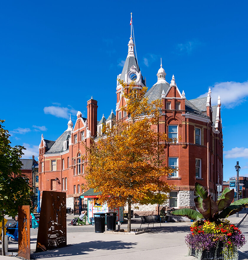 A historic red brick building with a tall clock tower stands under a bright blue sky, embodying strong roots. Autumn trees surround the scene. In the foreground, theres a modern art sculpture and a planter with colorful flowers. People walk nearby.