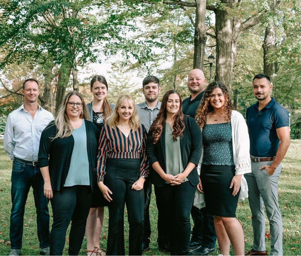 A group of nine Strong Roots employees stands together outdoors on a grassy area with trees in the background, symbolizing strong roots. They smile and pose casually, wearing a mix of professional and casual clothing. The scene is bright and sunny.