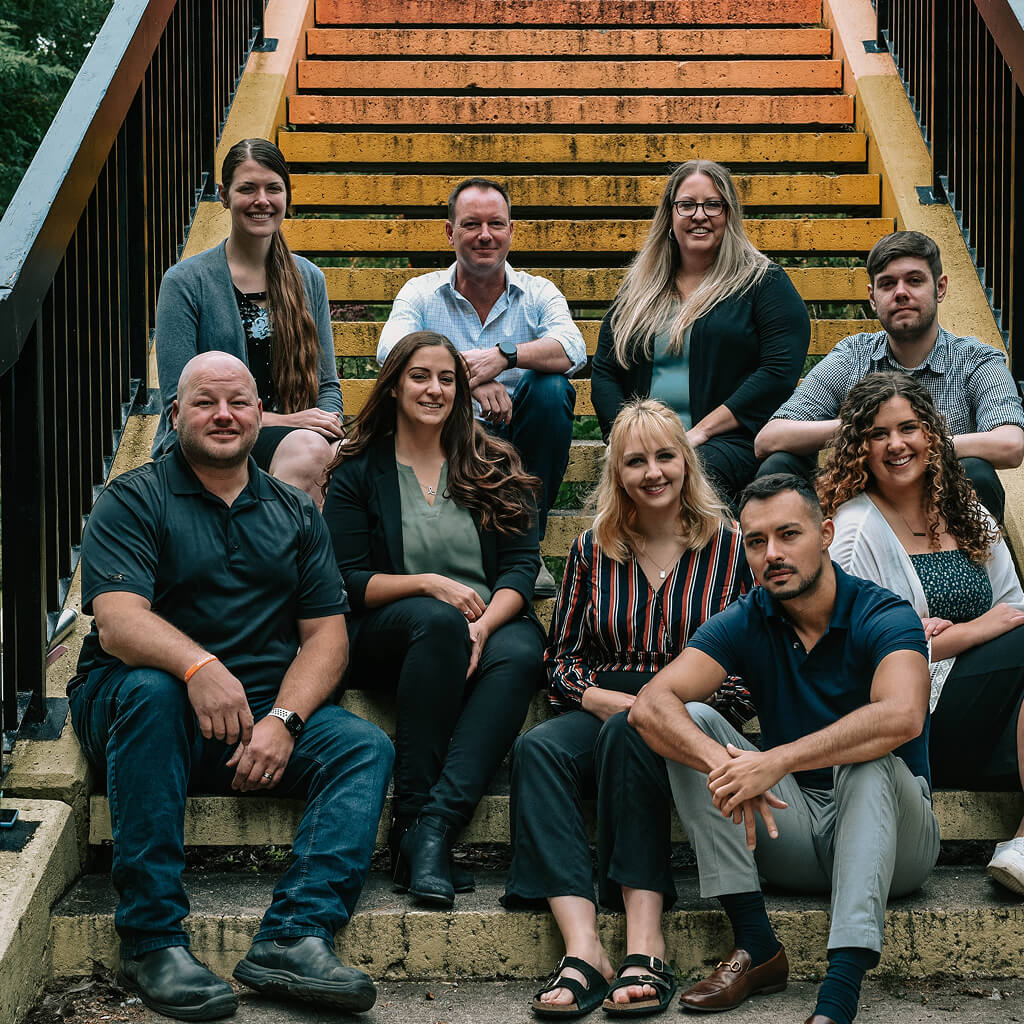 A group of nine Strong Roots employees, diverse in appearance, gather on outdoor stairs, embodying strong roots. Dressed in casual and business casual attire, they are united by their foundation. The steps and railing glimpse through warm sunlight, highlighting a shared connection.