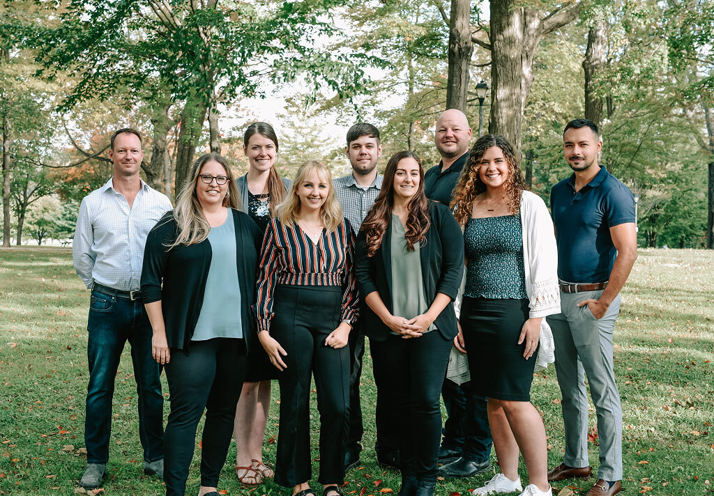 A group of nine Strong Roots employees stands outside on a grassy area with trees in the background, embodying strong roots. Dressed in casual and business casual attire, they are smiling for the photo. The setting suggests a sunny day in a park or outdoor space.