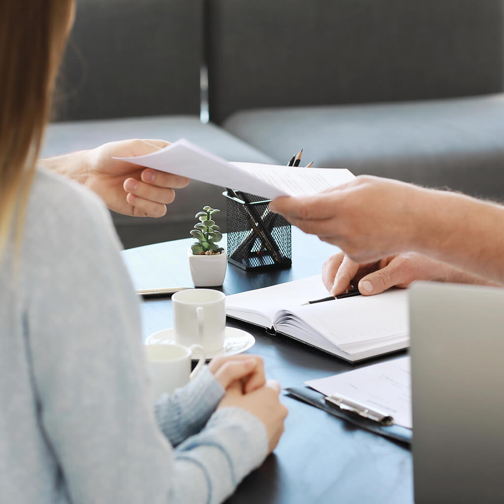 Two people seated at a table are exchanging documents. Among the items on the table—a laptop, notebook, and pens—sits a small potted plant symbolizing strong roots. The setting exudes a casual office vibe with its coffee cup and clipboard adding to the ambiance.