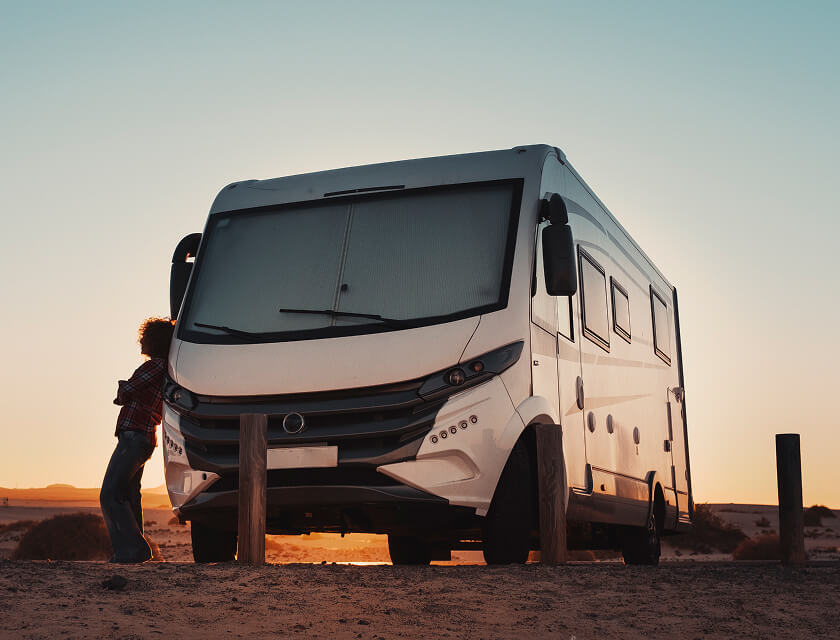 A large white motorhome, covered by auto insurance, is parked on a sandy terrain at sunset. A person leans against it, gazing at the sky. Wooden posts are nearby, and the horizon is visible in the background.