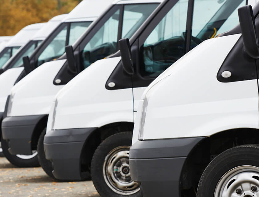 A row of white delivery vans, covered under comprehensive commercial insurance, are parked closely together. The focus is on the front sides of the vehicles, highlighting mirrors and part of the wheels. In the background, a few autumn trees stand serenely.