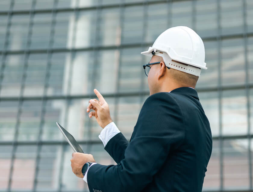 A man in a suit and hard hat holds a tablet, gesturing confidently. Standing before a modern glass building, he seems deeply engaged in planning or inspection—perhaps assessing the structures needs for commercial insurance.