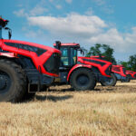 Four red tractors are lined up in a field under a blue sky with scattered clouds, embodying the essence of reliable farm insurance. The ground is covered in dry, harvested straw, while trees stand visible in the distance.