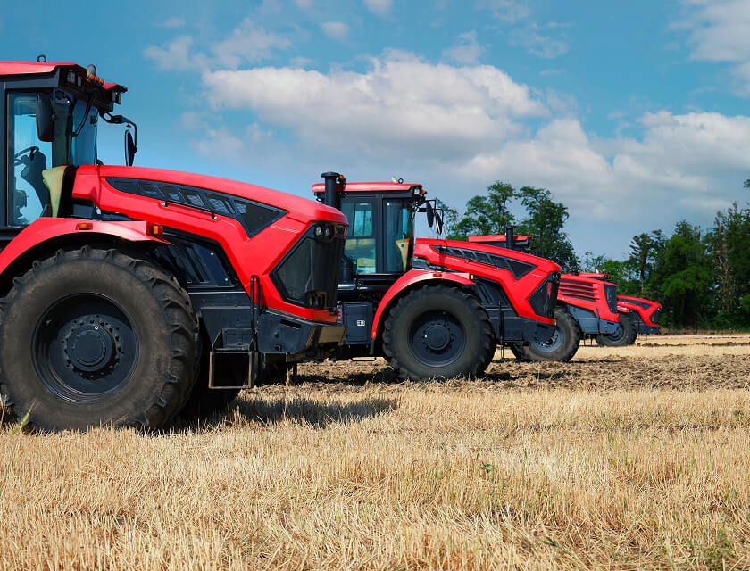 Four red tractors are lined up in a field under a blue sky with scattered clouds, embodying the essence of reliable farm insurance. The ground is covered in dry, harvested straw, while trees stand visible in the distance.