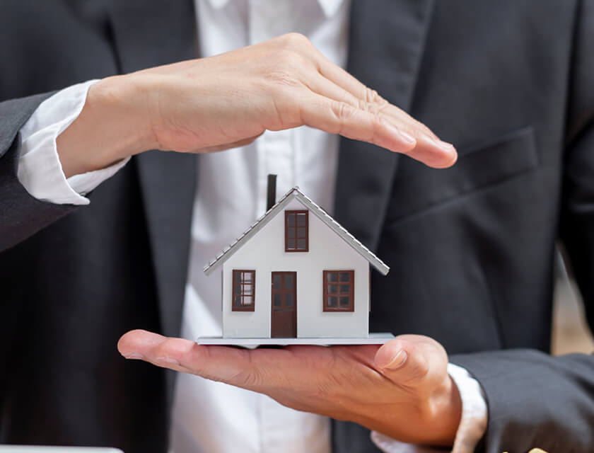 A person in a suit holds a model house, symbolizing the protective embrace of residential insurance. One hand cradles it below, while the other hovers above. The house is white with brown windows and a brown door, representing safety and shelter.