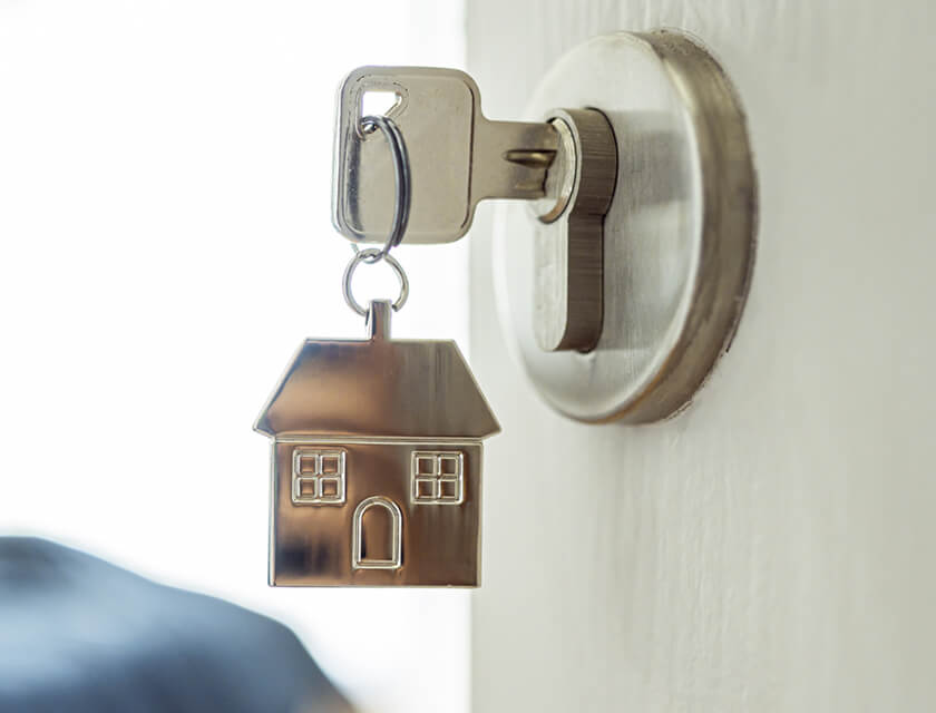 A silver house-shaped keychain, symbolizing residential insurance, is attached to a key in the door lock. The design features windows and a door, hinting at protection. The background is blurred, adding an air of mystery.