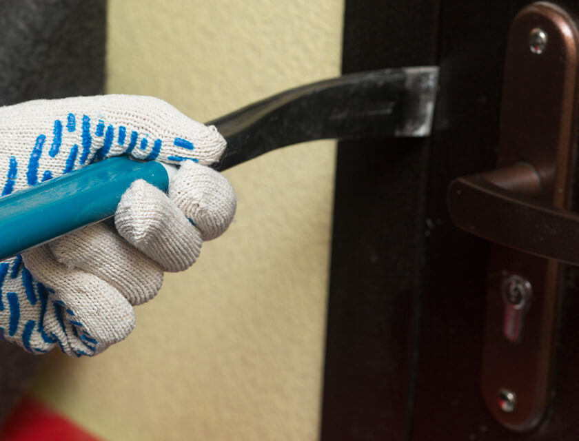 A gloved hand grips a crowbar, attempting to pry open a door. The door, secured with a metal handle, resists as the crowbar is wedged between the frame—a reminder of the importance of residential insurance for unexpected incidents.