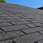 Close-up of a roof with gray asphalt shingles, arranged in a neat pattern as part of effective risk prevention. The clear blue sky forms the backdrop, while some debris is visible on the shingles.