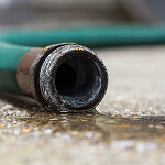 Close-up of a worn green garden hose with water trickling out onto a wet concrete surface. The pooling water is a gentle reminder to employ risk prevention resources to avoid unexpected slip hazards in the future.