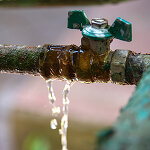 A leaking green metal water valve with water streaming from the connection point suggests a need for risk prevention. The valve is partially rusted, and the background is blurred, emphasizing the urgency in managing resources effectively to prevent further damage.