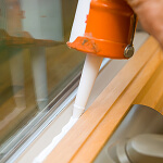 A hand holds an orange caulking gun, applying white sealant to the corner between a window frame and the wall, utilizing resources for risk prevention. The sealant is applied smoothly along the wooden frame, with the glass reflecting light.