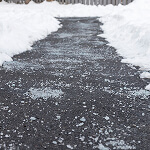 A shoveled path, a testament to risk prevention, is clear of snow and bordered by towering snowbanks. The dark asphalt peeks through, sprinkled with salt granules for ice melting. This path leads safely to a wooden fence in the background.