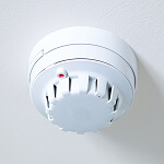 A white smoke detector, a vital risk prevention resource, is mounted on the ceiling. The round device features a central body with vent-like gaps around its perimeter and a small red indicator light visible on the side.