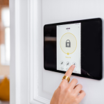 A hand with a gold ring is touching a wall-mounted smart home security tablet displaying a "Secure" status with a padlock icon, and options for different zones like "Living Room" and "Outdoor."