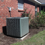 An outdoor air conditioning unit, or HVAC system, sits on a concrete pad next to a red brick house with a white-trimmed window, surrounded by green grass and some bushes with pink flowers.