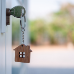 A silver key with a house-shaped wooden keychain is inserted into a lock, with a blurry background of greenery and a light-colored building.