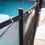 A close-up view of a black mesh pool safety fence with a dark post, with the blurry blue water of a swimming pool visible in the background under clear skies.