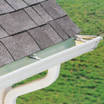 A clean, white eavestrough and downspout attached to the side of a house with dark grey asphalt shingle roofing, against a blurry green grass background.