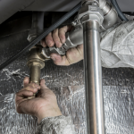 A person's hands are shown working on connecting or repairing a complex network of metal pipes, possibly part of a plumbing or HVAC system.