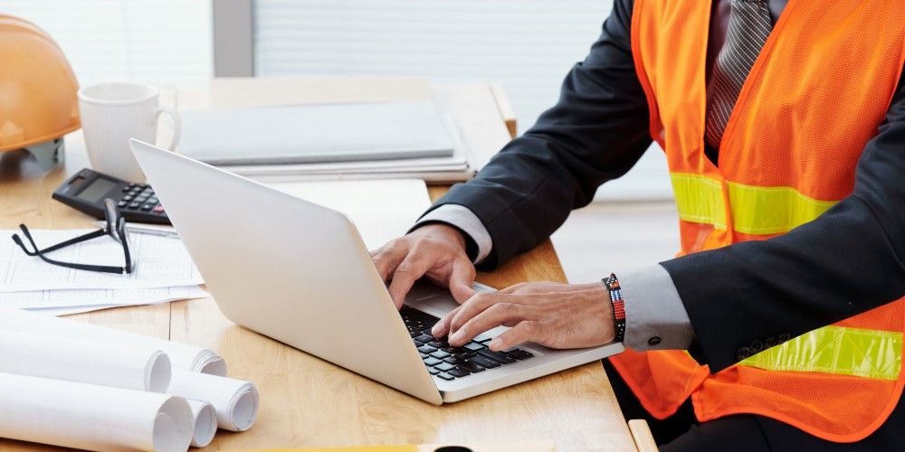 Unrecognizable man in neon safety vest and business suit sitting at desk and using laptop