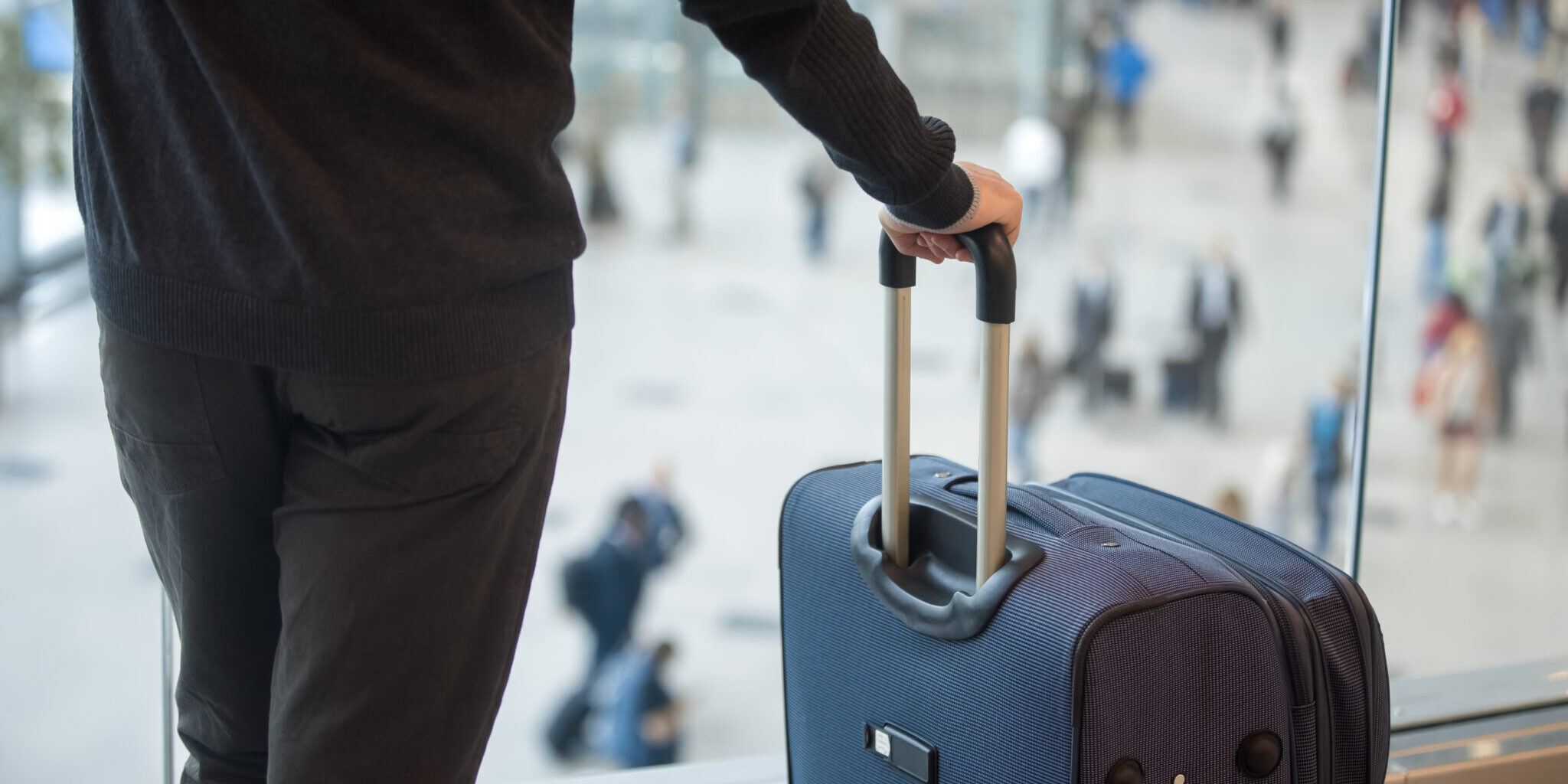 Young man waiting for flight, standing in modern airport terminal with crowd on background, holding handle of luggage bag, wearing casual clothes, rear view, close up