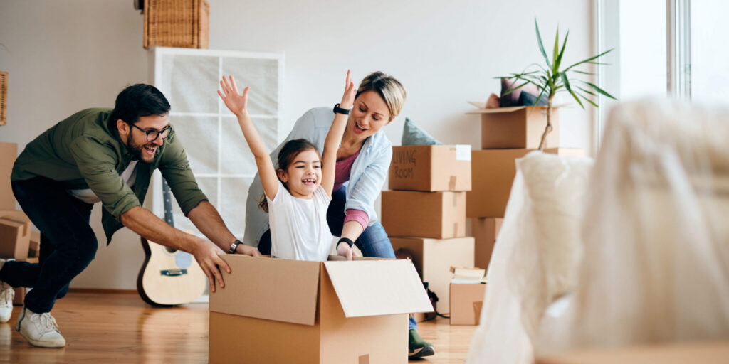 Playful parents pushing their daughter cardboard box and having fun at new apartment.
